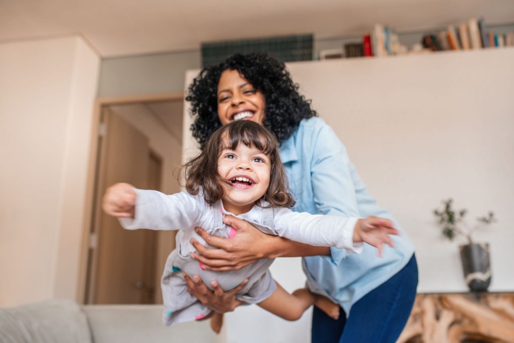 Foster mother holding a little girl in the air in the living room as if the girl were flying like an airplane